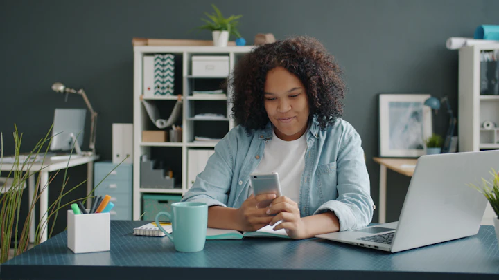 Mulher sorridente usando o celular em uma mesa de trabalho com notebook, em casa